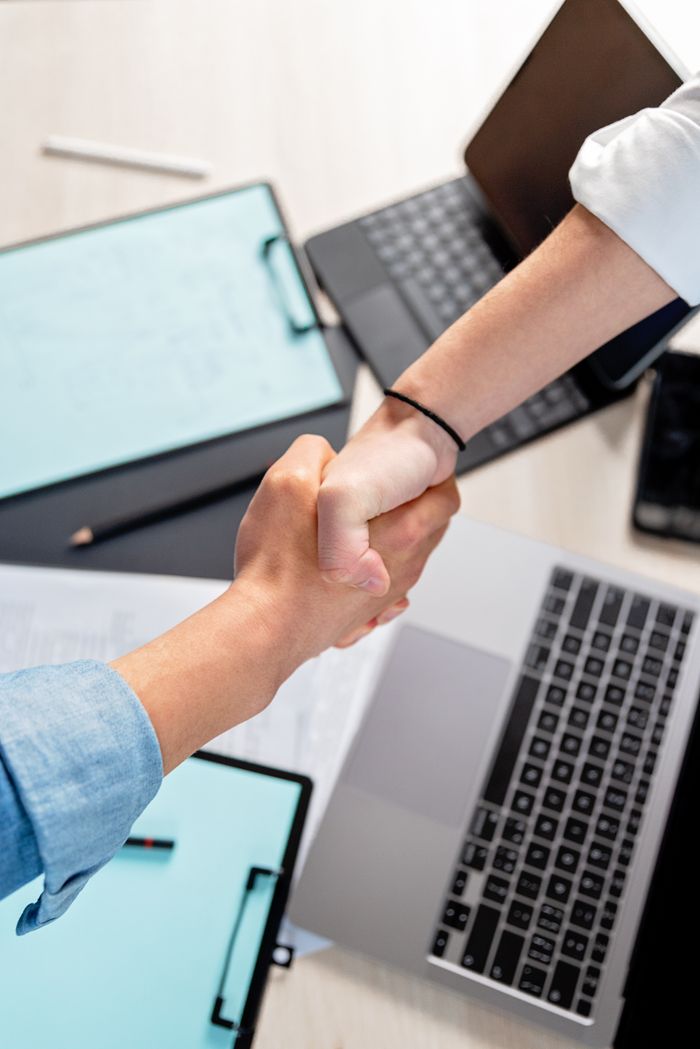 portfolio-04 Two people shaking hands over laptops and documents in an office, symbolizing a business deal.